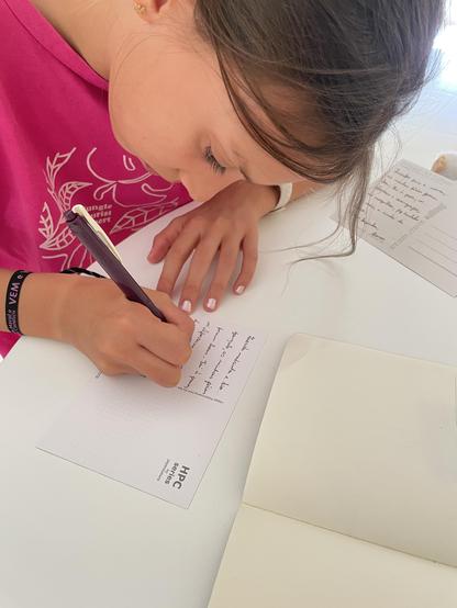 A young girl in a pink shirt writes carefully on the back of a postcard at a white table.