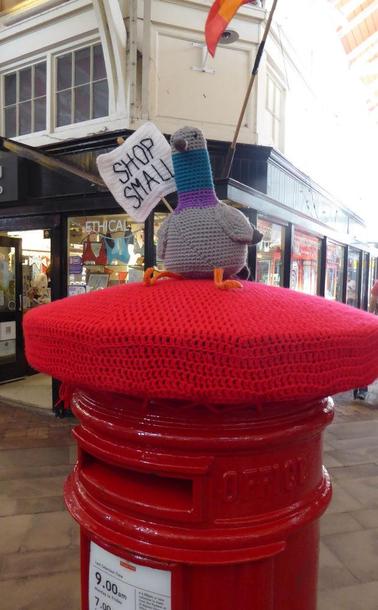 Red postbox with red, knitted cover on top, atop of which is a knitted pigeon holding a sign that says 'SHOP SMALL'.