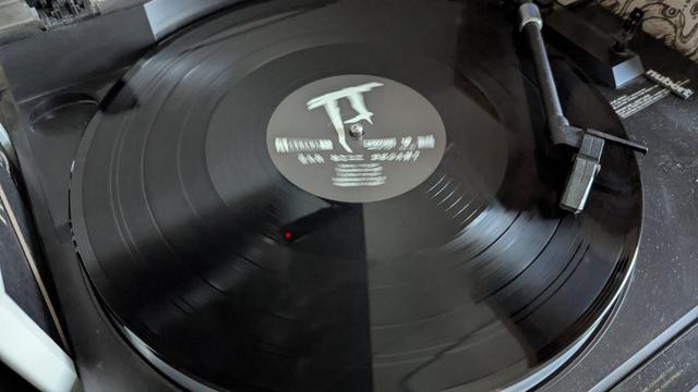 Nahaufnahme einer Schallplatte, die sich auf einem Plattenteller dreht, mit einer über der Platte positionierten Abtastnadel. Das Etikett auf der Schallplatte ist teilweise sichtbar und zeigt ein abstraktes Design und Text.