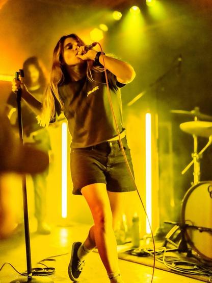 A female singer performs on stage, holding a microphone, with long hair and wearing a dark t-shirt and shorts. The scene is illuminated with yellow lighting, creating a vibrant atmosphere. In the background, band members and musical instruments can be seen.