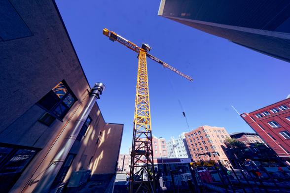 In the center of the frame a large crane with a yellow space frame mast and boom looks smaller than it really is with the boom pointing towards the lower right with the hook halfway down,  to the left there is a beige building in dark shade close to us,  at the upper right another building close to us sticks up in a sharp corner also in the shade,  and towards the bottom a distance row of brick and pastel buildings is brightly lit and there is roughly a triangle of blue sky in the middle.