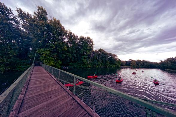 A wood bridge with diagonally oriented planks and green mesh metal sides goes from filling the whole frame at the bottom to about 20% left  on the vertical center line with a dark triangle of water on the left and to the right a water area with ripples and I think 6 kayaks each with one paddler,  deciduous trees fill a triangle from the upper left to about the right 1/3 then there is a thin strip of trees that gets a little wider just before the right of the frame,  above there are probably str…