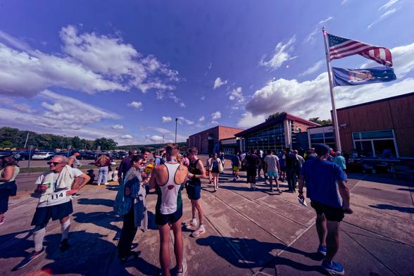 Older and younger people wearing various sorts of athletic clothes mill around on the concrete Plaza in front of a school building on the right with a flagpole with an American flag with a New York flag under it,  in the distance can be seen parked cars,  a row of trees,  and a blue sky with mostly big cumulus clouds but some higher altitude clouds too.