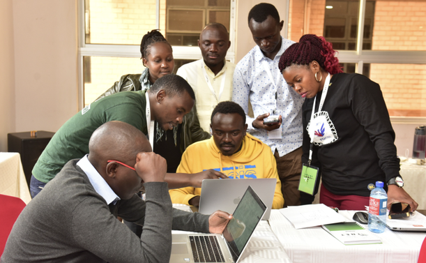 A group of seven people, four men and three women, are gathered around two laptops in a collaborative workspace. They appear engaged and focused, discussing something on the screens. One man in a yellow hoodie is seated and using the laptop, while others stand or lean in to contribute. Notebooks, pens, and a water bottle are on the table. The setting is bright with large windows in the background. Source: APC.org – Financing digital equity: WSIS+20 Taskforce, Credit: Kenyan National School of Community Networks. Photo: TandaNet