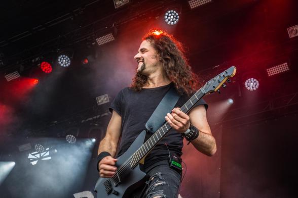 Das Bild zeigt einen Musiker auf einer Bühne, der eine schwarze Elektrogitarre spielt. Er hat lange, wellige Haare und trägt ein schwarzes T-Shirt und abgerissene Jeans. Der Musiker lächelt und scheint sich in seiner Performance zu befinden. Die Bühne ist mit verschiedenen Lichtern beleuchtet, darunter rote und weiße Scheinwerfer, die eine dramatische Atmosphäre schaffen. Im Hintergrund sind einige Lichter und eine Kulisse zu sehen, die auf eine Live-Auftrittszene hinweisen. Der Musiker hat ein…