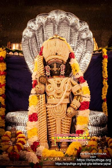 Idol of Hindu deity Krishna in a "chain mail" of pearls with gold undergird, standing with a matching dome-shaped crown and a similarly clad staff in right hand. A thick string of yellow, white, and red roses around Krishna. A large coiled 7-headed silver cobra ["shesha"] behind Krishna with the snake's head forming an umbrella over Krishna. Dark background.