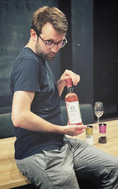 Ein Mann mit kurzen braunen Haaren und einer schwarzen Brille sitzt in einem Theater-Probenraum gespielt lässig auf der Kante eines Esstisches und hält aufreizend eine volle Flasche Roséwein in die Kamera.