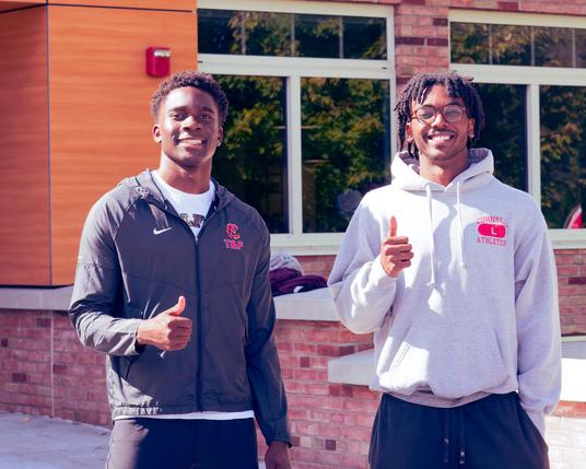 Two black men give the thumbs up gesture with their right (your left) hands,  the one on your left has a grey Cornell hoodie with a Nike logo and short hair,  the one on your right has glasses and short dreadlocks and a white Cornell sports hoodie.  Behind them is a short brick wall,  wood panel column with a fire alarm and two windows divided by a thin section of brick wall.