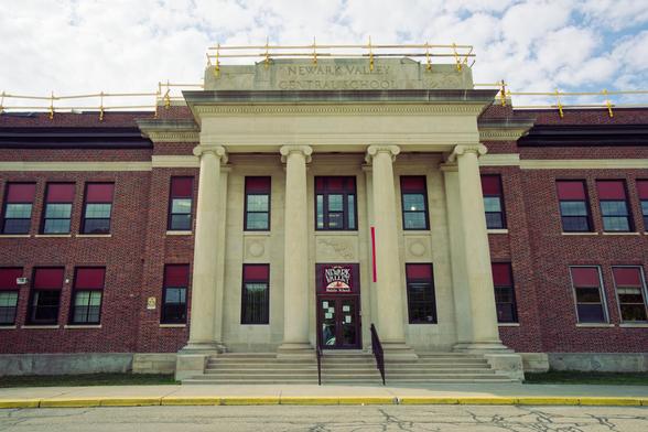 Two-story brick building with marble-looking classical columns in the center with a sign at top that reads NEWARK VALLEY CENTRAL SCHOOL and a set of yellow scaffolding around the roof.