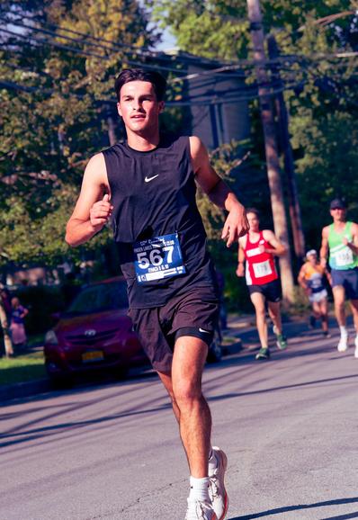 In front a man with a black Nike tank top and short hair with a blue bib that says 567 in white is ahead of several other runners in the middle of a road with a red compact car in the shadows behind him,  power lines, trees and a bit of a Victorian house.