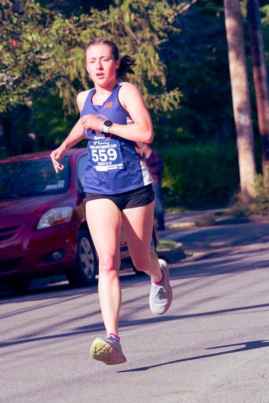 A woman with her hair tied back running fast down the road with both feet suspended and a blue tank top,  the number 559 I think an Apple Watch and Black shorts.  Behind her a bit of a red car, evergreen tree,  and telephone pole.