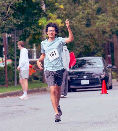Man with black curly hair is flying with both feet off the ground and his left thumb (your right up) while a volunteer in shorts and a white sweatshirt looks off the left of the frame and a barely visible volunteer holds a red flag to stop a black car by a safety cone.