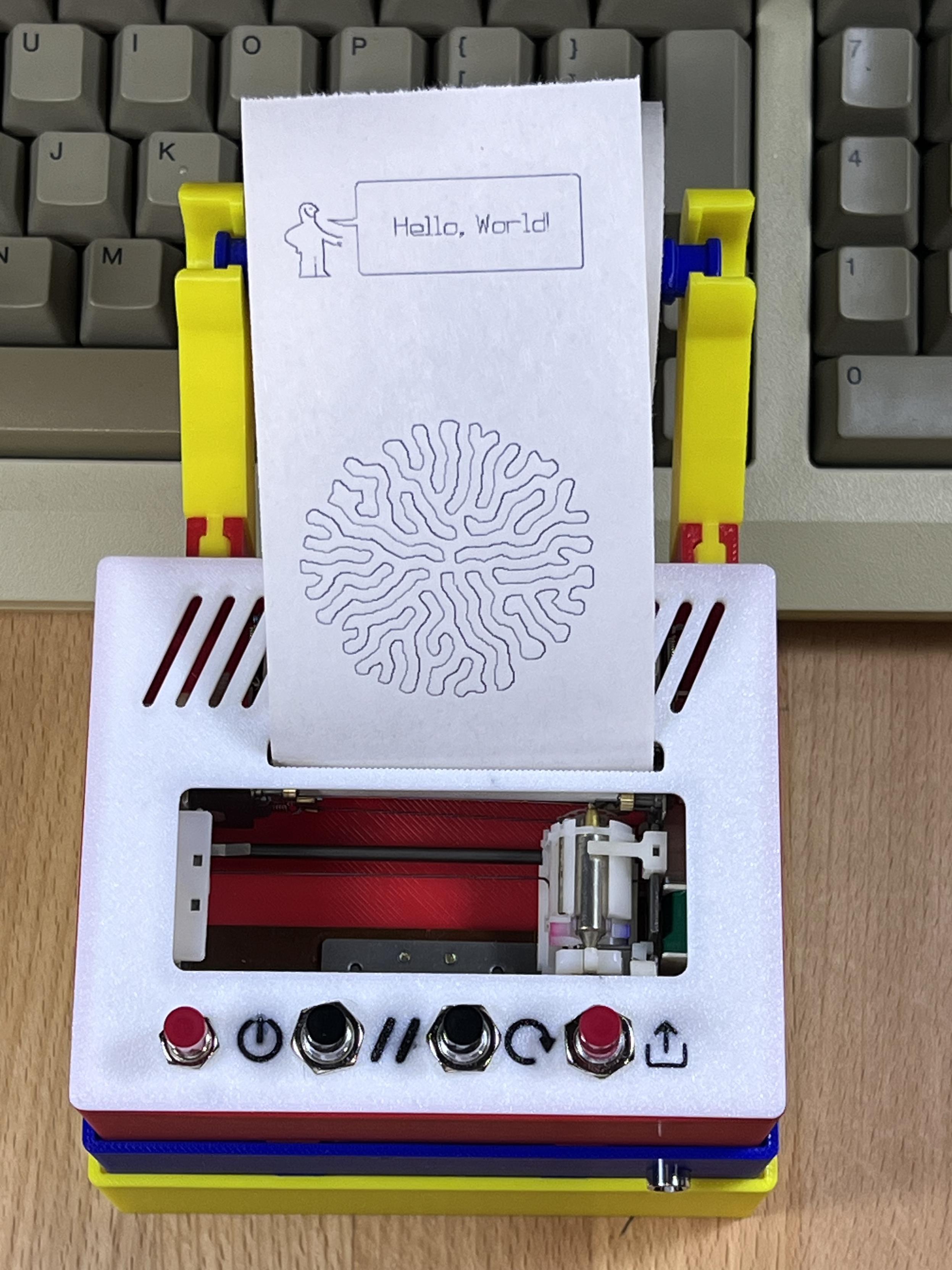Top view of a tiny pen plotter housed in plastic boxes. A white top with buttons and paper coming out show the text Hello, World and a differential growth pattern drawn on it. Top view of a tiny pen plotter housed in plastic boxes. A white top with buttons and paper coming out show the text Hello, World and a differential growth pattern drawn on it.