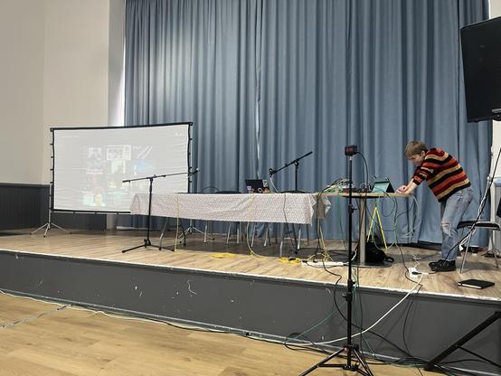 A – not yet very interesting – view of the stage at the Alpaca conference, showing a projection screen, a long table, a couple of mic stands and a blue curtain behind. The AV engineer is busy poking at a laptop on the table.