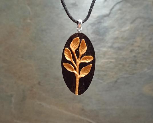 A sprig of six tiny leaves sprouting from a single central twig, carved in golden boxwood, mounted to an upright oval of black ebony wood. Silver bail, and a black cord keeping it suspended. With an out of focus grey slate background.