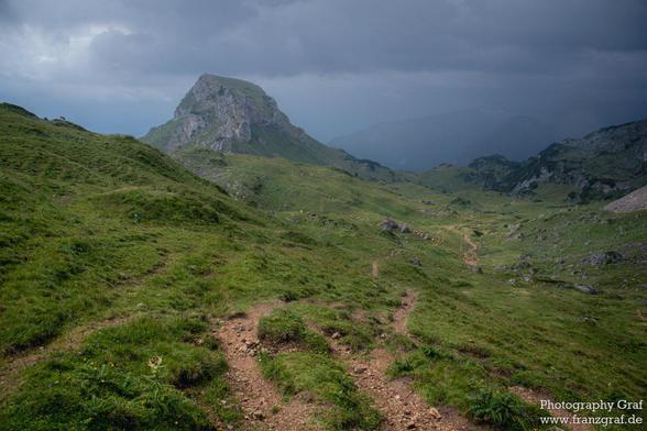 A picturesque scene of a grassy hill with a majestic mountain in the background is depicted in this image. The grassy hill is vibrant green, contrasting beautifully with the grey mountain towering in the distance. The sky above is dotted with fluffy clouds, adding a touch of serenity to the landscape. This outdoor setting evokes a sense of peace and tranquility, perfect for nature lovers and outdoor enthusiasts. The highland terrain is rugged yet inviting, with a hint of wilderness and adventur…