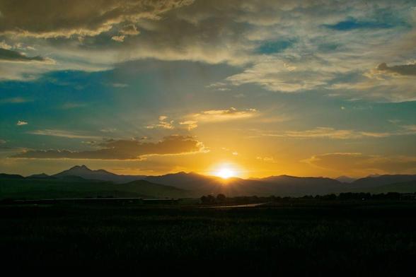 Sunset over Longmont, Colorado, with the sun dipping behind silhouetted mountain ridges. The sky glows in golden and orange tones, with soft clouds scattering the light. The Twin Peaks of Longs Peak and Mount Meeker are faintly visible to the left.