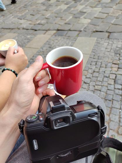 Ruka na ulici drží červený hrnek s kávou, před hrnkem na koleni odpočívá černá filmová zrcadlovka.
