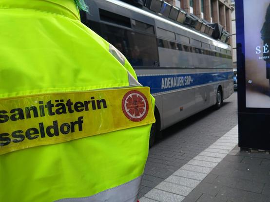 Im Vordergrund Links im Bild der Rücken einer Demosanitäterin mit dem Rückenschild Demosanitäter Düsseldorf drauf stehen, und Rechts im Bild der Bus Adenauer SRP+ 