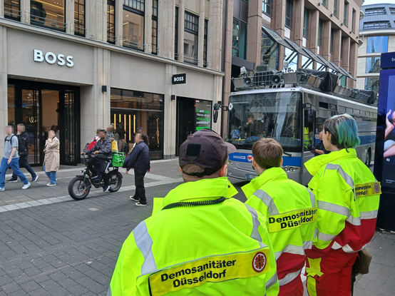Im Vordergrund 3 Demosanis die auf denn Vorbeifahrenden Bus gucken.