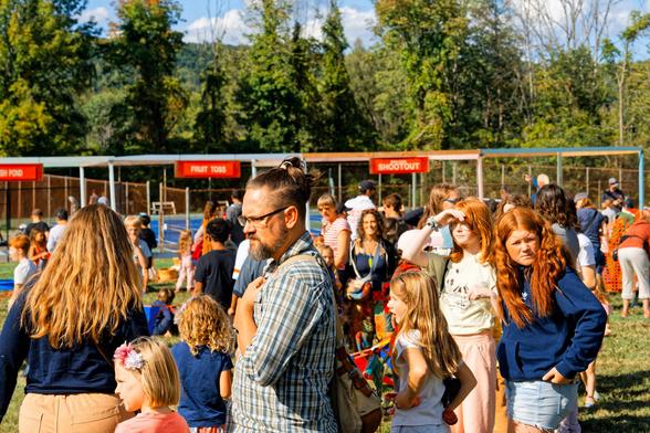 A large crowd of men, women and children are milling around on an Early fall day in front of a line of carnival games with red signs and behind that a hedgerow and far past that a rolling hill and a blue sky with cumulus clouds