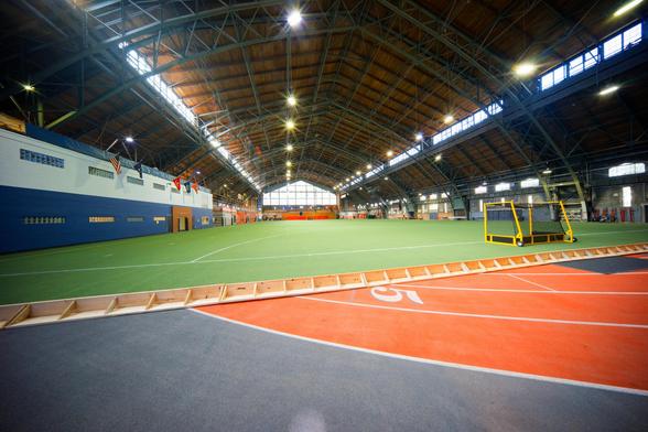 A wide-angle shot of the inside of a large clear span structure with an indoor running track that is interrupted by a short plywood barrier and past that a field of green artificial turf with a yellow goal structure with a black net, and to the left a building inside the building with a blue first story and white second story.