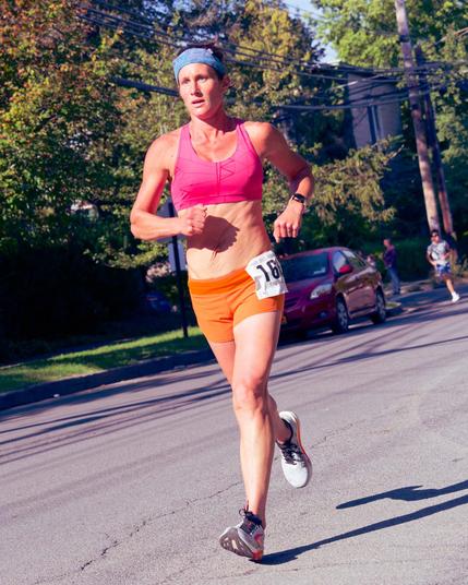 Woman running with her left (your right) foot striking at the heel and the other behind her up in the air,  she's wearing yellow shirts and a pink top that esxposes the midriff and a blue headband,  behind are some street trees in a subdued green, another runner in the distance and a lot of power lines.