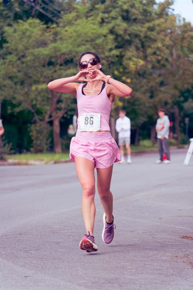 A woman wearing a pink tank top and pink shorts and purple shoes and dark shades has both feet suspended at the moment she makes a heart sign with her two hands right in front of her face;  behind her can be seen two volunteers managing traffic and spreading deciduous trees.