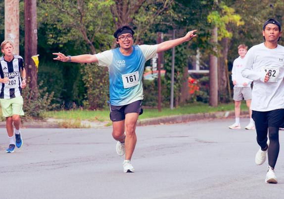 In the center of the frame a man with the number 161 and a gradient-colored shirt that is blue in the middle to greenish white on the (short sleeves) and (yout) bottom right has his arms spread wide.  He's got a headband, glasses and white shoes.  202 is partially visible on the left and he has a shirt with wide black and white stripes that look like a european soccer uniform,  another male runner is partially cisible on the right and has a white sweatshirt and black running tighets and white s…