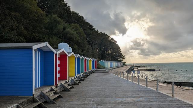 Brightly-coloured beach huts line the promenade, with dark, mature trees looming behind. The sun sinks into tumbling clouds above a choppy sea