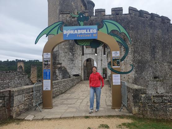Une photo d'Anormally qui se tient debout sous le panneau d'entrée du festival. On peut y voir un grand dessin de dragon et y lire Bonabulles. Derrière ce panneau on voit une partie du château dans lequel se déroule le festival.