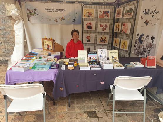 Photo de Anormally au stand des trois canards. Il y a deux tables colorées recouvertes de nos livres et de badges pour la palestine. Derrière des affiches de nos éditions et une expo de "juste un bisou"