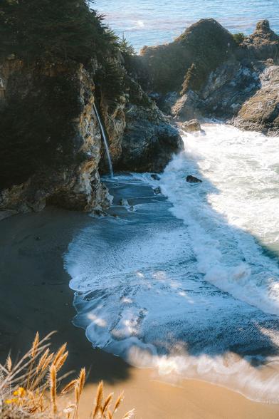 A secluded beach with gentle waves, a small waterfall, and greenery on rocky cliffs in the background.