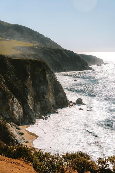 A panoramic view of a coastline with steep cliffs, gentle waves, and a sandy beach under bright sunlight.
