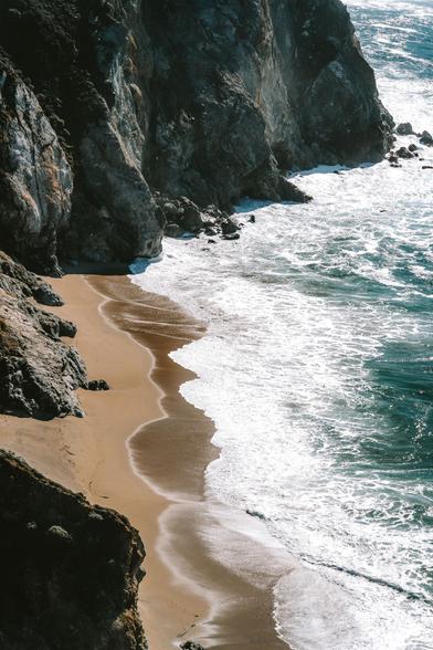 A sandy beach bordered by steep cliffs, with waves lapping at the shore and rocks scattered along the coastline.