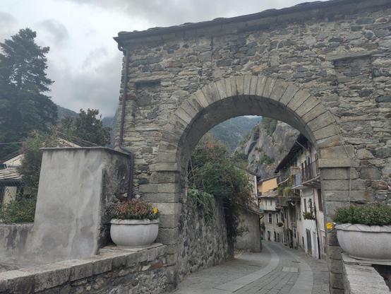 Arcus lapideus in Via Francigena prope Pontem Sancti Martini, in Pedemonte, vicum stratamque saxeam et aedificia rustica cum vasis florum et viriditate ascendente cingens.