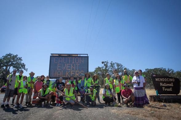 Group of people in safety vests pose near a “Caution Event Walkers” sign and Mendocino National Forest sign under a clear blue sky.