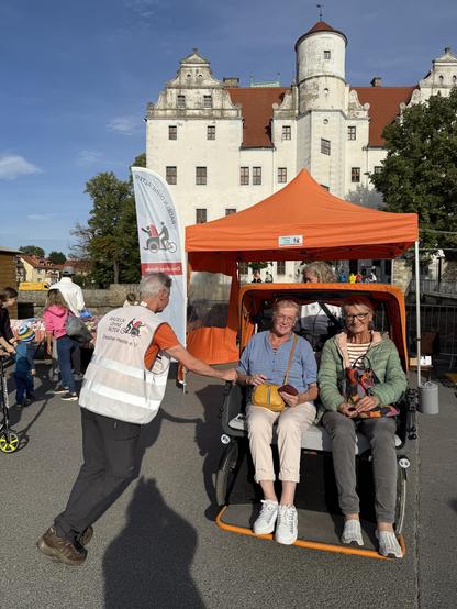 Zwei Passagierinnen in der Rikscha und Pilot Jürgen mit weißer Warnweste und unserem Vereinsaufdruck vor dem Schloss