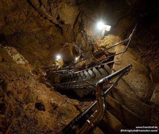 A dimly lit cave is depicted in this image, showcasing a staircase made of metal that leads further into the mysterious depths. The walls of the cave are a dark shade of brown, adding to the eerie atmosphere of the scene. The staircase stands out against the backdrop, serving as a pathway for exploration into the unknown. The image captures the sense of adventure and curiosity that comes with delving into the underground world. The ground beneath the staircase is rough and textured, hinting at …