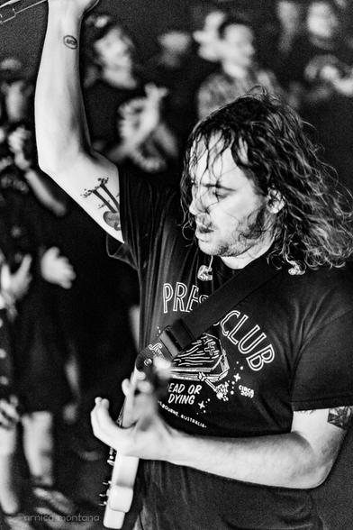A musician plays guitar on stage, wearing a black t-shirt with the words "PRESS CLUB" and an illustration. They exhibit energetic movement, with long, curly hair and a visible tattoo on their arm. The audience is blurred in the background.