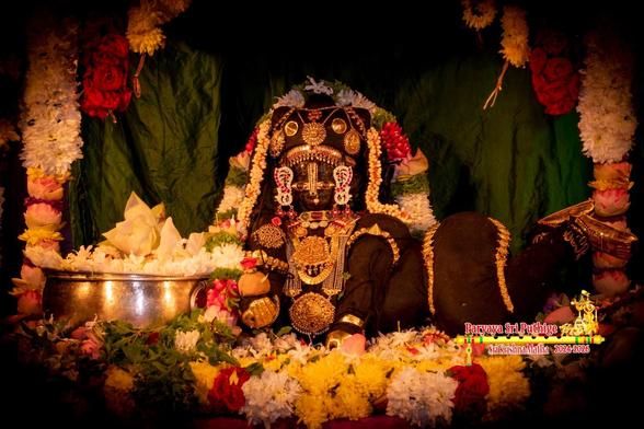 Closeup of idol of Hindu deity Krishna dressed in black as a baby crawling in a bed of assorted loose flowers; wearing lots of gold ornaments and head turned to us; a thin string of white flowers and a thick string of white and red flowers (some green holy basil leaves) around Krishna. A silver bowl filled with white flowers in front of Krishna to his right. A string of assorted flowers including lotuses and roses forming an arch. Dark green cloth background.