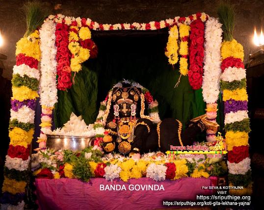 Idol of Hindu deity Krishna dressed in black as a baby crawling in a bed of assorted loose flowers; wearing lots of gold ornaments and head turned to us; a thin string of white flowers and a thick string of white and red flowers (some green holy basil leaves) around Krishna. A silver bowl filled with white flowers in front of Krishna to his right. Colorful strings of assorted flowers (including lotuses and roses) forming an arch over Krishna. Pink (magenta) cloth on the platform for the whole assembly. Dark green cloth background.