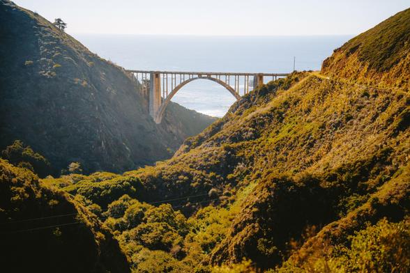 A tall bridge arches over a green valley, connecting two hilly landscapes with ocean in the background.