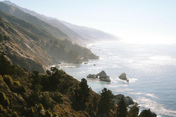 Mist-covered coastline with rocky outcrops and distant mountains, under a clear sky.