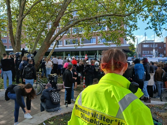 Eine Demosanitäterin steht auf erhöter position und guckt auf Linke sowie Rechte Demonstrantinnen und eine Behelmte Polizeikette.
