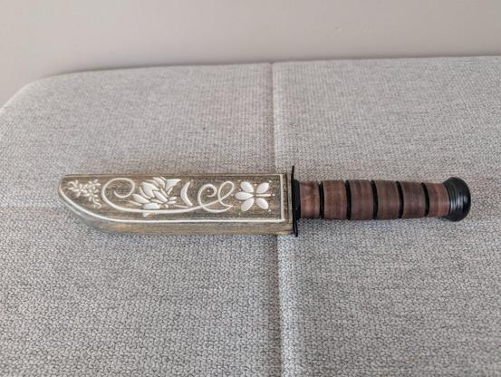 A K-Bar knife with a hand carved scabbard. The scabbard is stained to look weathered and has a floral pattern carved into it. Where the grooves and carvings are, there is white enamel paint.