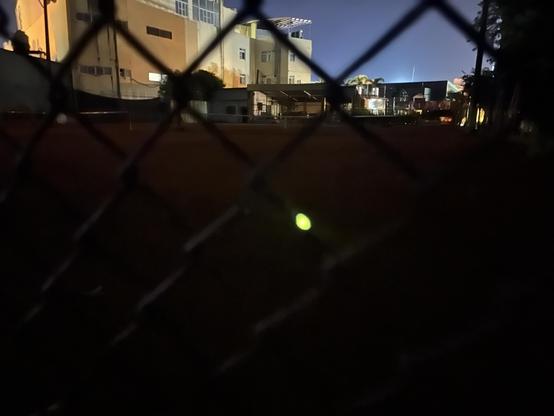 A blurry nighttime long exposure photo of the chain-link fence of a tennis court with buildings and the night sky in the background.  In the foreground on the chain-link fence is the bright yellow-green blob of light of a firefly.