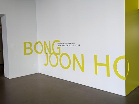 White wall with BONG JOON HO in yellow, and in black,
DIRECTOR'S INSPIRATION 
LA INSPIRACION DEL DIRECTOR