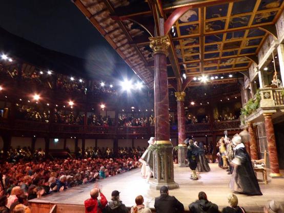 Side view of Elizabethan covered stage within circular timbered theatre. Lights shining on stage as cast take their bows. Night.