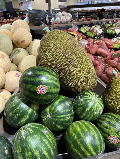 on top of a pile of cantaloupes and small watermelons sits a very large durian. Behind are bags of potatoes. 

I don’t recall ever seeing such an exotic fruit in this part of the county (mostly deep red) 